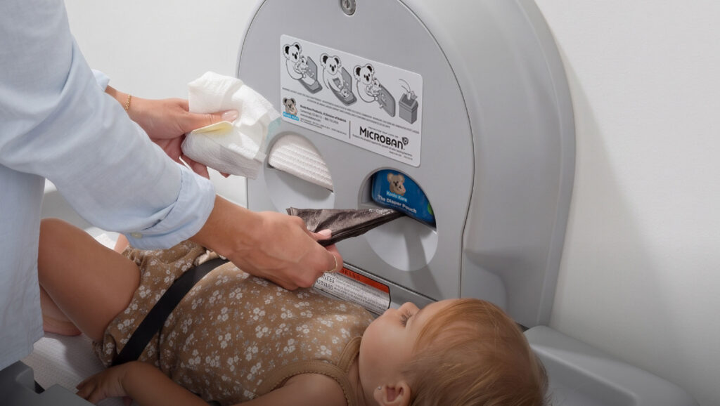 Close-up of a parent changing a toddler on a wall-mounted Koala Kare baby changing station, pulling a dark disposable diaper bag from the station’s integrated diaper disposal bag dispenser for The Diaper Pouch cartridge.
